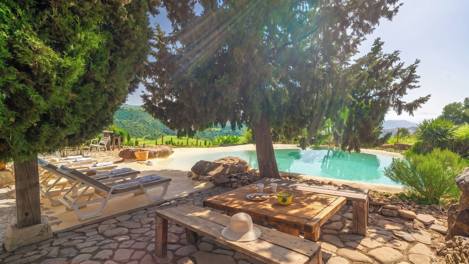 A poolside area with lounge chairs, a wooden table, and a view of a mountain landscape.