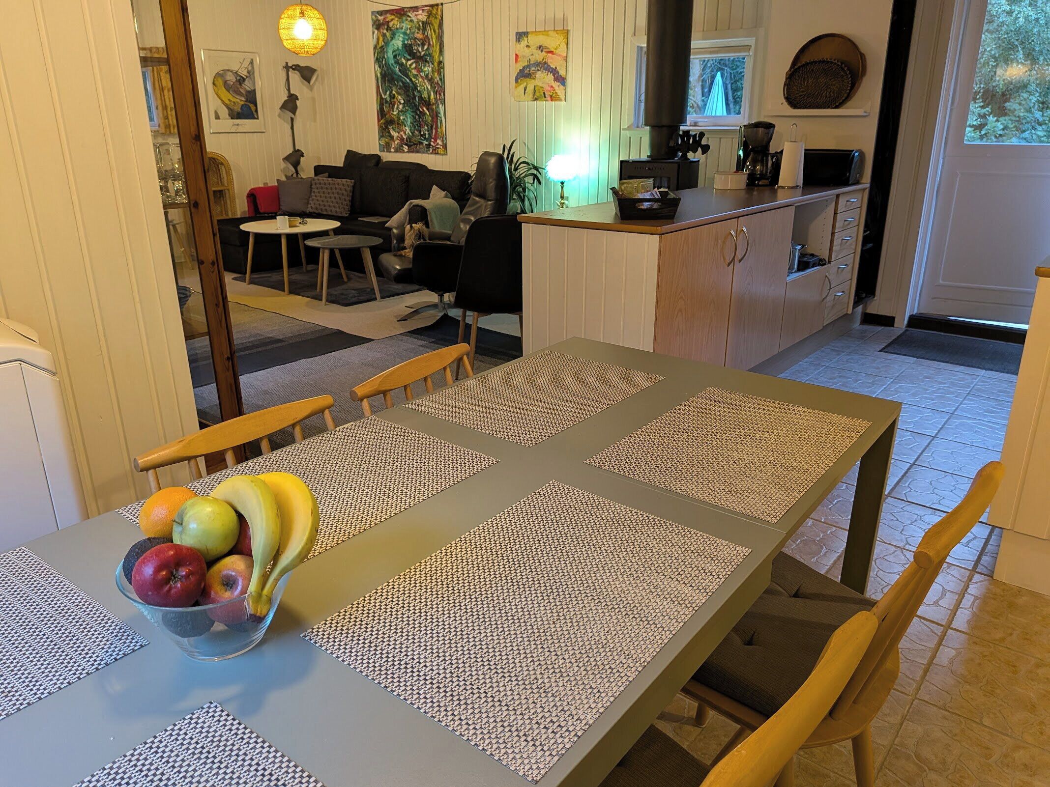 Een eethoek met een tafel gedekt voor twee personen, een schaal fruit en een keuken op de achtergrond.
