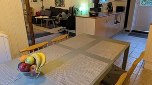 Un coin repas avec une table dressée pour deux personnes, un saladier de fruits et une cuisine en arrière-plan.