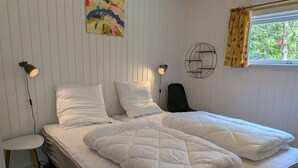 Un lit avec une literie blanche, une lampe de chevet, une chaise, un tableau décoratif accroché au mur et une fenêtre avec des rideaux.