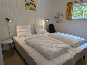 Ein Bett mit weißer Bettwäsche, eine Nachttischlampe, ein Sessel, ein Wandbild und ein Fenster mit Vorhängen.