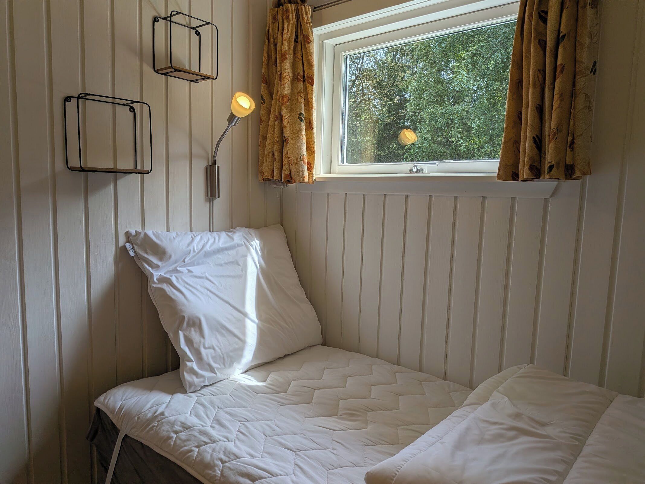 Ein Einzelbett mit weißer Steppdecke, einer Nachttischlampe und einem Fenster mit Vorhängen.