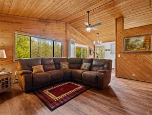 Ein Holzblockhaus-Interieur mit einem Ecksofa, einem roten und schwarzen Teppich, einem Deckenventilator und einem gerahmten Landschaftsbild.