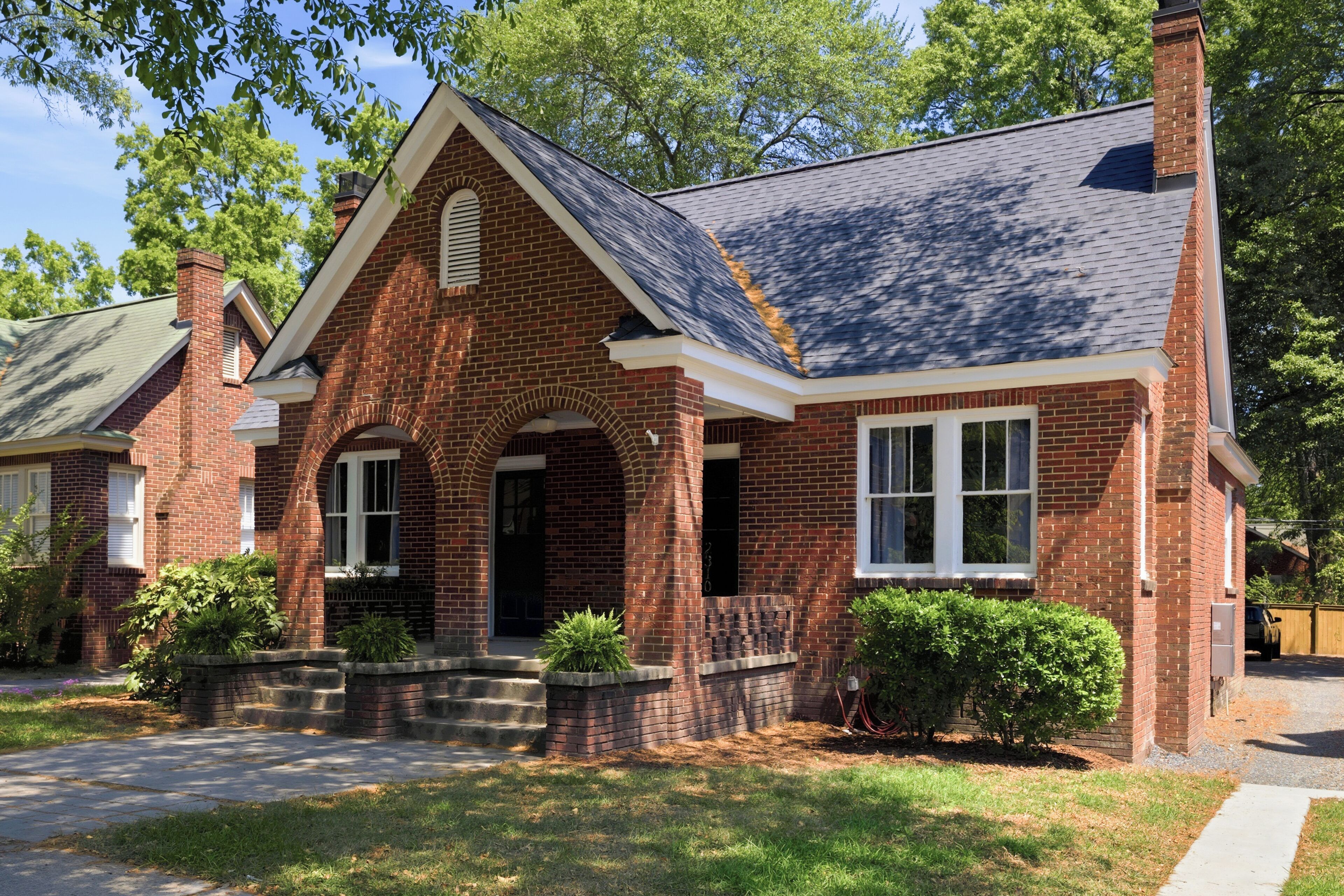 A brick house with a gabled roof, a front entrance with steps, and a small front yard with shrubs.