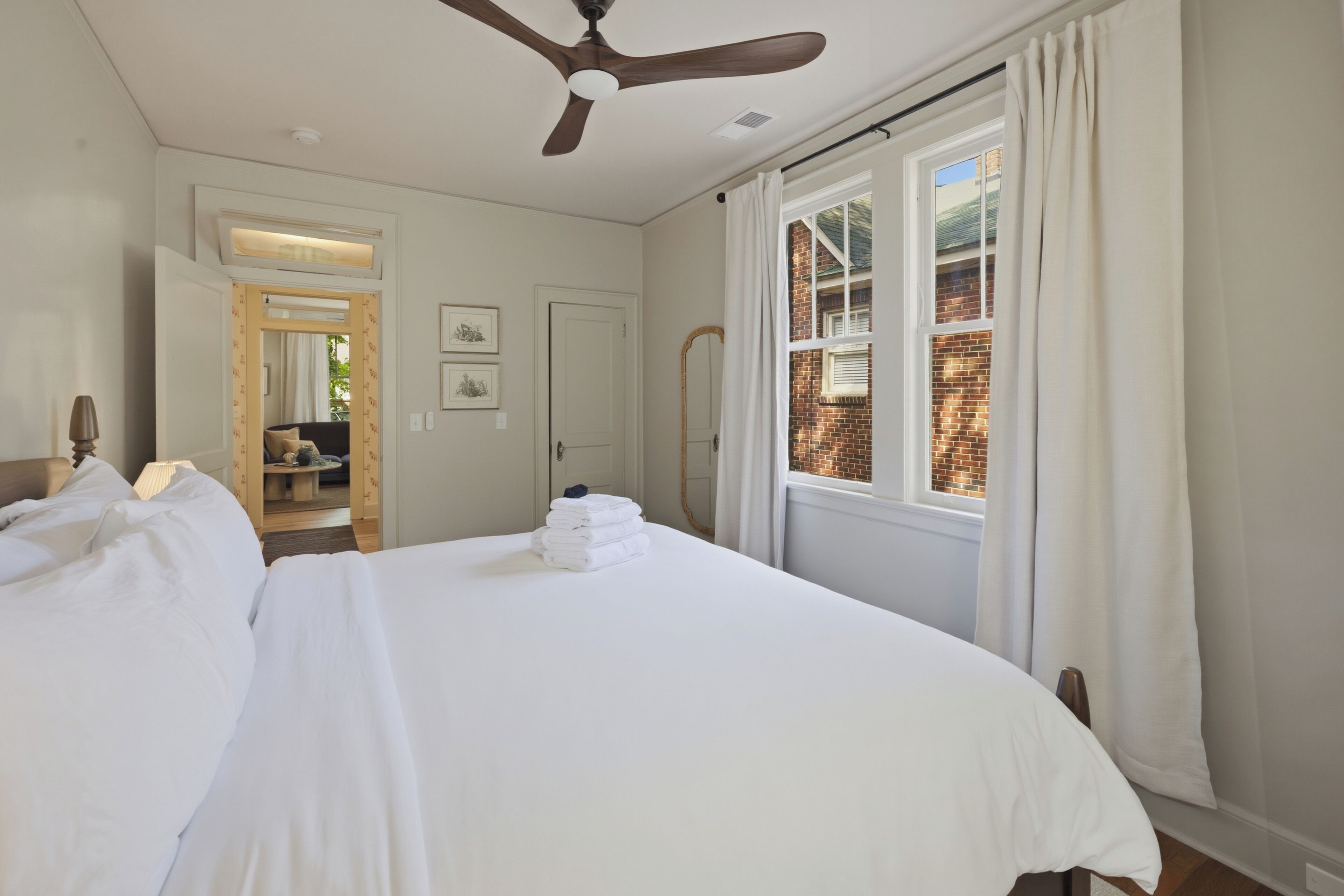 A bedroom with a large bed, a ceiling fan, a window with curtains, and a view of a brick building.