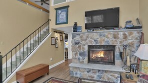 A living room with a stone fireplace, a television mounted above it, and a staircase with a glass railing. - Midlake Retreat Waterfront 3BR Penthouse, Lake Club Included, Pool, Ski & Golf (Lake Harmony)