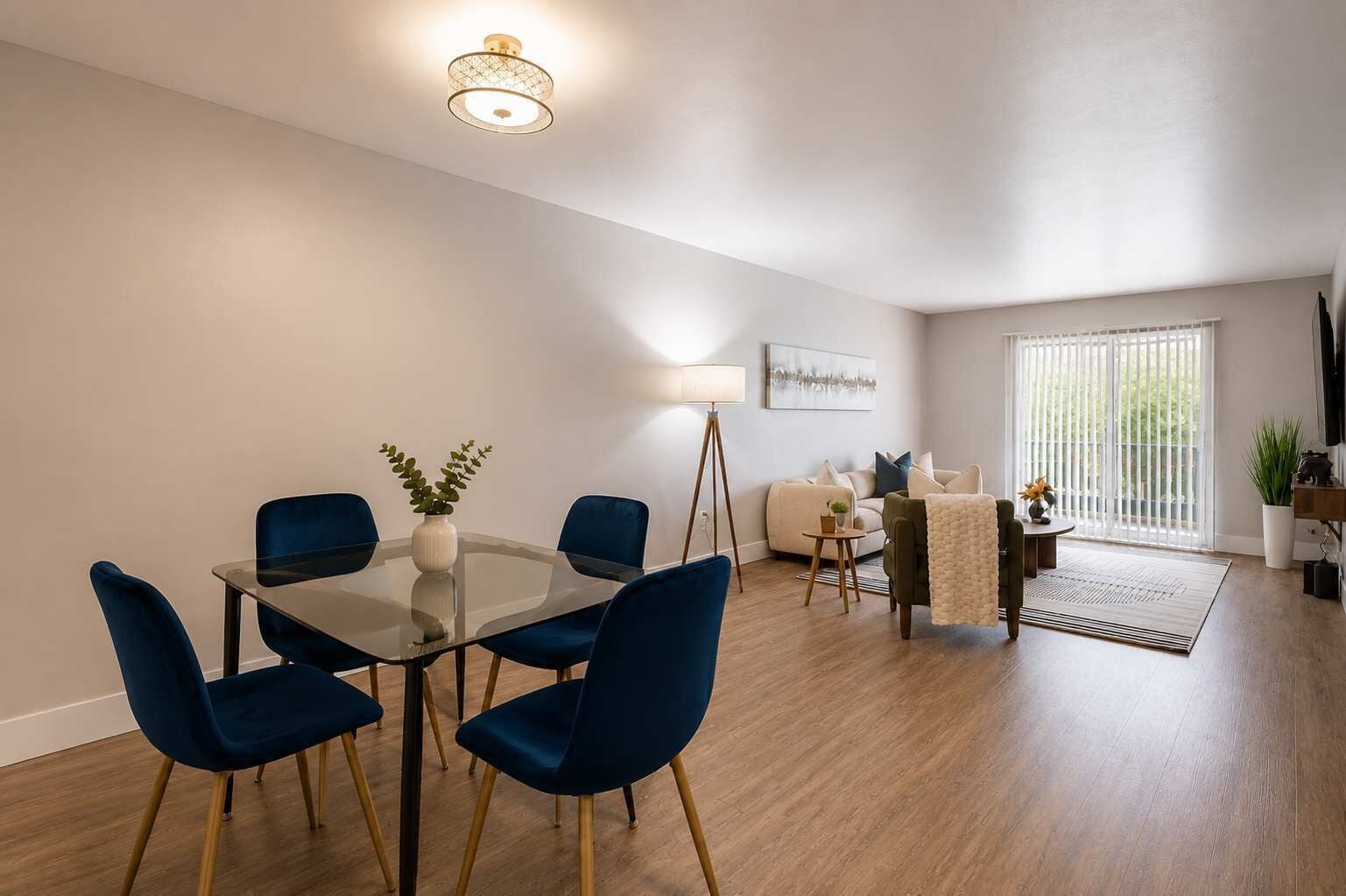 Un salon moderne avec une table à manger en verre, des chaises bleues, un canapé, une lampe sur pied et une grande fenêtre avec des stores.