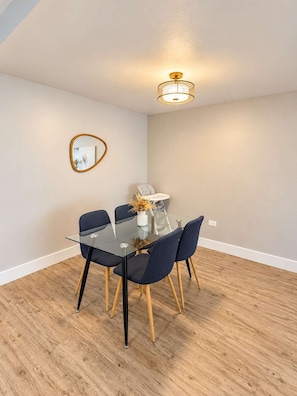 Un coin repas avec une table en verre, des chaises foncées, un vase contenant des fleurs séchées, un miroir au mur et un luminaire suspendu au plafond.