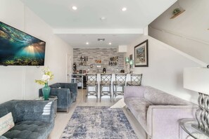 A modern living room with a large TV, two sofas, a coffee table, and a kitchen area with bar stools. - The Cottages Collection - 10 Oceanfront Suites and 2 BR Penthouse (East End)