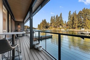 Een terras met een tafel en stoelen, uitkijkend op een meer met bomen en gebouwen op de achtergrond.