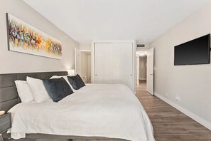 Een slaapkamer met een bed, een televisie, een schilderij en een deur die naar een andere kamer leidt.