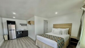 Un dormitorio con cama, cabecera de madera, mesita de noche y una zona de cocina integrada con refrigerador y lavabo.