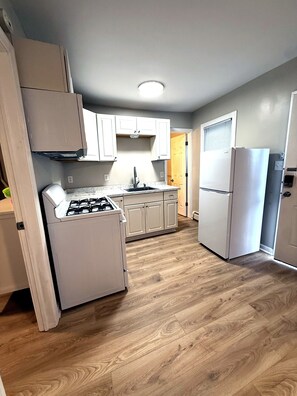 A kitchen with white cabinets, a gas stove, a refrigerator, and a wooden floor. - A Slice of Seaside Charm — Comfortable 2-Bedroom Cottage - 2 blocks to beach (Seaside Heights)