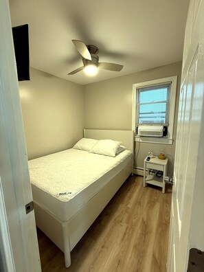A small, neatly arranged bedroom with a single bed, a ceiling fan, a window with blinds, and a bedside table with a lamp and a clock. - A Slice of Seaside Charm — Comfortable 2-Bedroom Cottage - 2 blocks to beach (Seaside Heights)