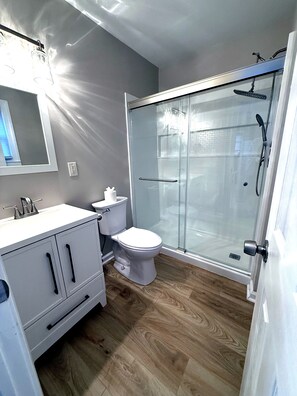 A modern bathroom with a glass shower enclosure, a white toilet, and a vanity with cabinets. - A Slice of Seaside Charm — Comfortable 2-Bedroom Cottage - 2 blocks to beach (Seaside Heights)