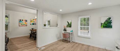 A spacious room with hardwood flooring, a desk with a chair, a window with blinds, and artwork on the walls.