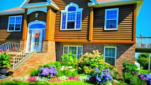 A two-story house with a front porch, a brick foundation, and a well-maintained garden. - 270° Stunning Ocean View & 4 Private Epic Gardens! (Terence Bay)