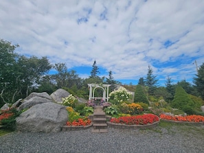 A garden with a central white arbor, surrounded by large rocks and colorful flower beds. - 270° Stunning Ocean View & 4 Private Epic Gardens! (Terence Bay)
