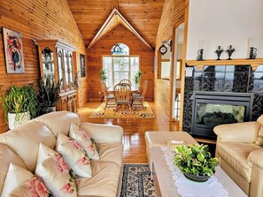 A cozy log cabin interior with a fireplace, wooden walls, a dining area, and a sofa. - 270° Stunning Ocean View & 4 Private Epic Gardens! (Terence Bay)