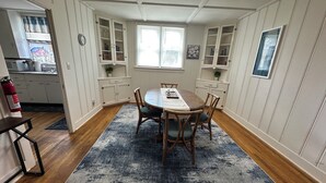 Un comedor con una mesa de madera, sillas y un tapete azul. Hay armarios blancos con puertas de vidrio, una ventana con persianas y un extintor en la pared.