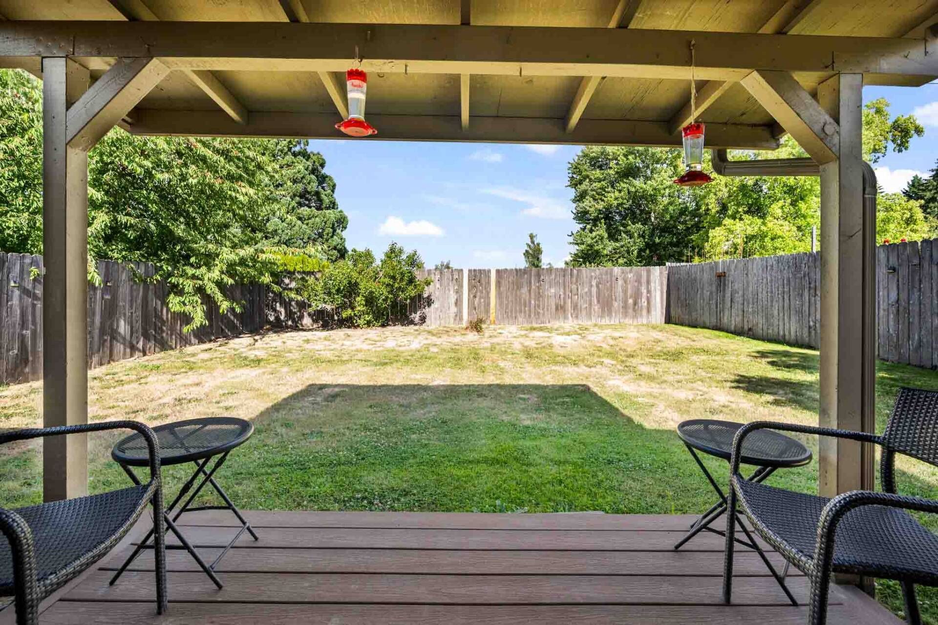 A covered patio with two chairs and a table, a wooden deck, and a fenced backyard.