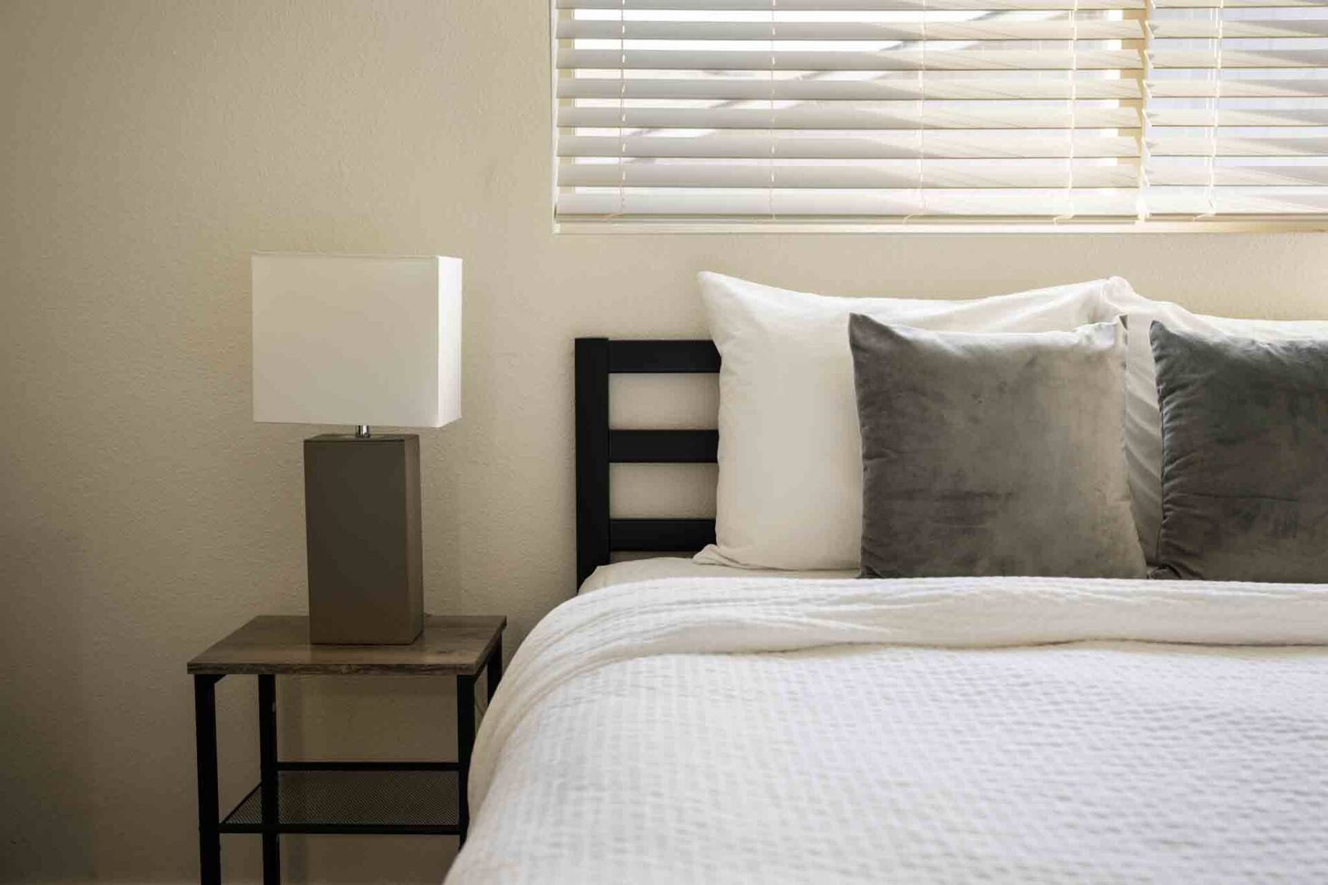 A neatly made bed with white bedding, a bedside table with a lamp, and a window with blinds.