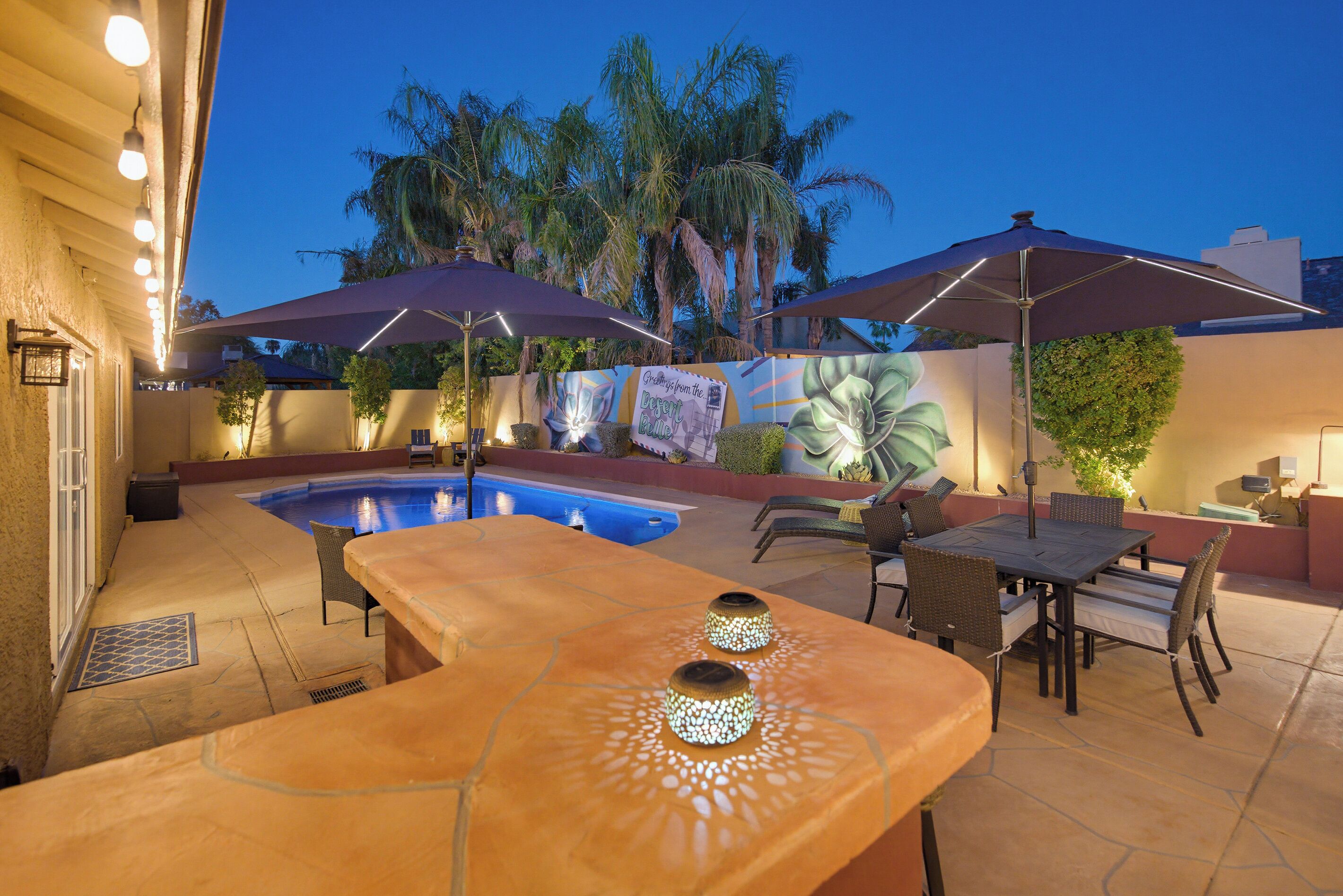 An outdoor pool area with a patio table and chairs, surrounded by palm trees and a mural.