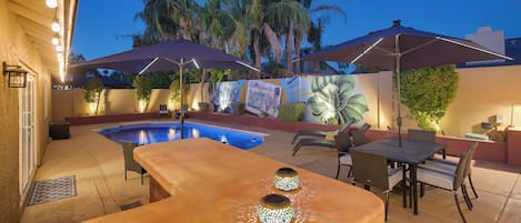 An outdoor pool area with a patio table and chairs, surrounded by palm trees and a mural.