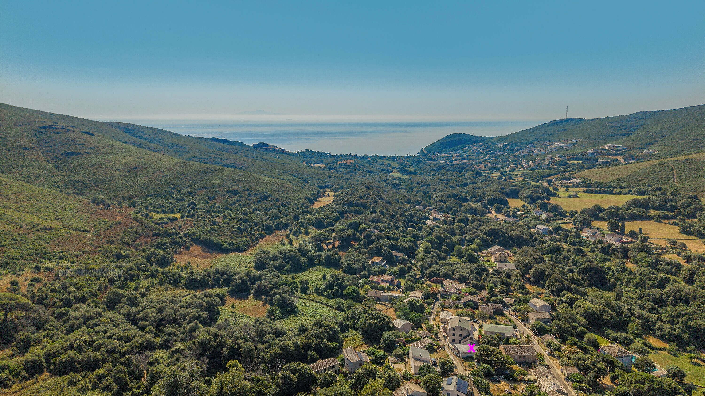 Una vista aerea di un borgo collinare con case sparse, cielo sereno e un corpo d'acqua in lontananza.
