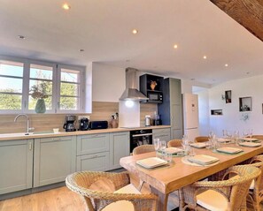 A modern kitchen with a dining area, featuring a long wooden table set for six, light-colored cabinetry, and a large window providing natural light. - A Stay in the Countryside - SAINTE COLOMBE (SAINTE COLOMBE)