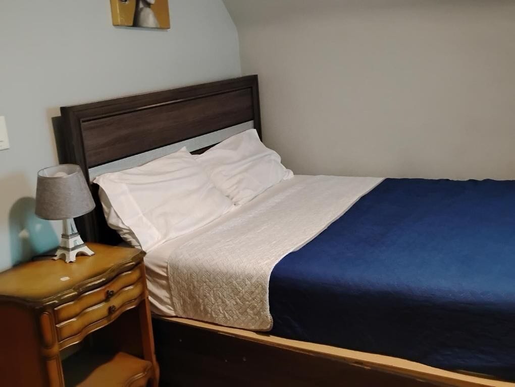 Een bed met houten hoofdeinde, een nachtkastje met lamp en een aan de muur hangend schilderij.