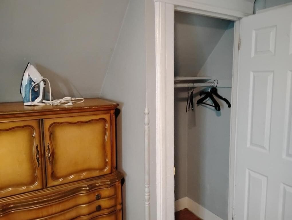 A wooden dresser with a white iron, a closet with hanging clothes, and a white door.