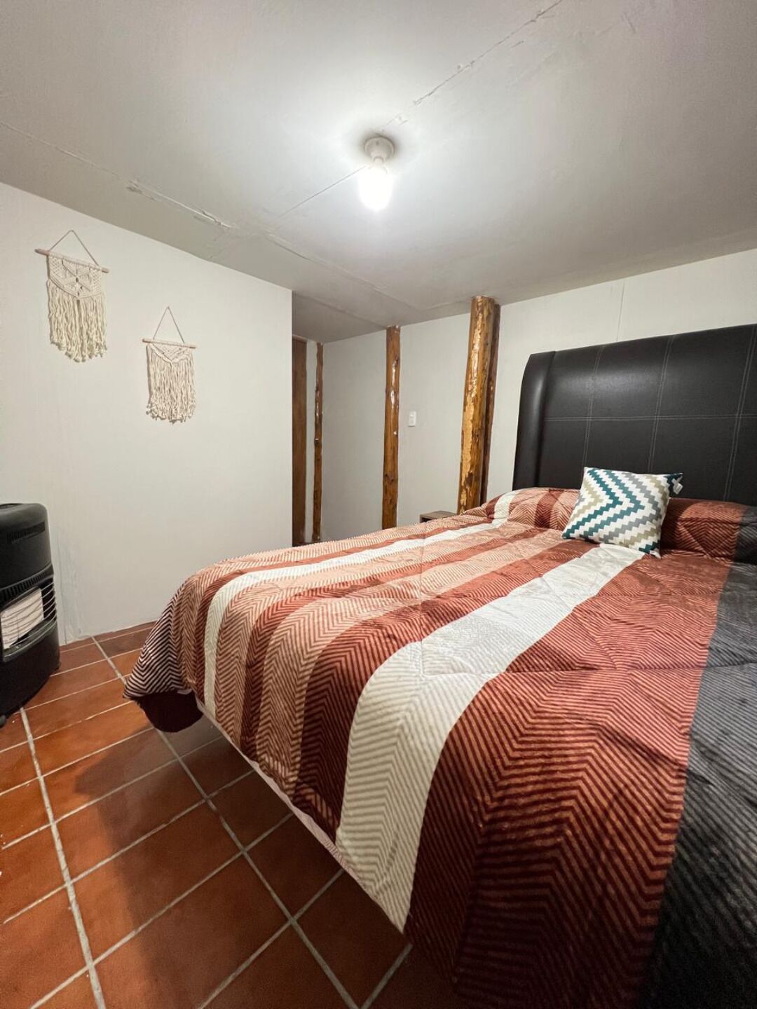 A bedroom with a bed, a black trash bin, a wall-mounted light fixture, and a ceiling with exposed wooden beams.