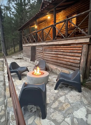 A log cabin with a stone patio, outdoor seating, and a fire pit.
