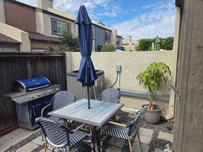 Un patio al aire libre con una parrilla, mesa, sillas y una sombrilla.