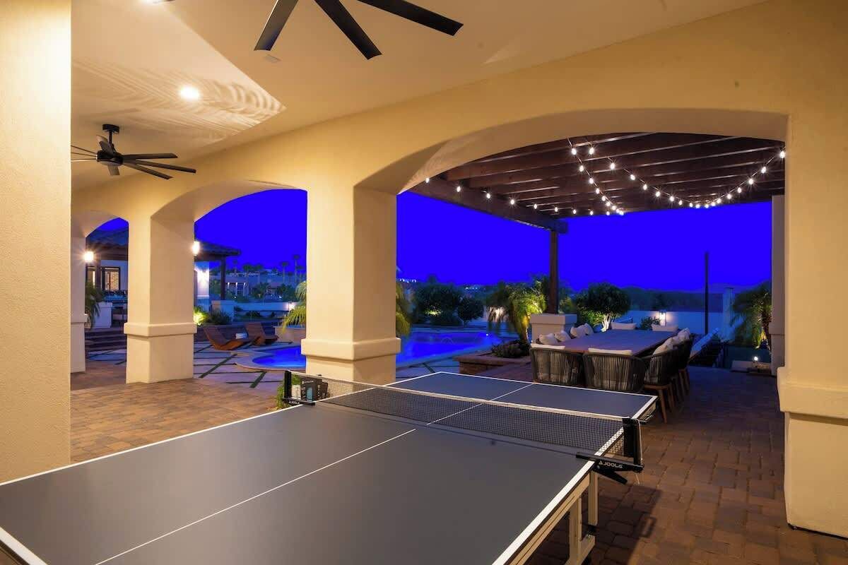An outdoor ping pong table with a covered seating area, a ceiling fan, and string lights.