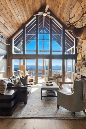 Ein geräumiges Wohnzimmer mit hoher Decke, großen Fenstern und einer Steinwand mit aufgehängtem Hirschkopf.