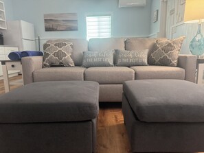 A living room with a grey sofa, matching ottomans, and decorative pillows with text. - BEACHSIDE TWO BEDROOM VILLA - PARTIAL OCEAN VIEW (Georgetown)