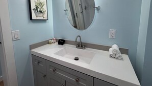 Une salle de bain avec un lavabo blanc, un miroir rond, un tableau encadré et une serviette posée sur le comptoir.