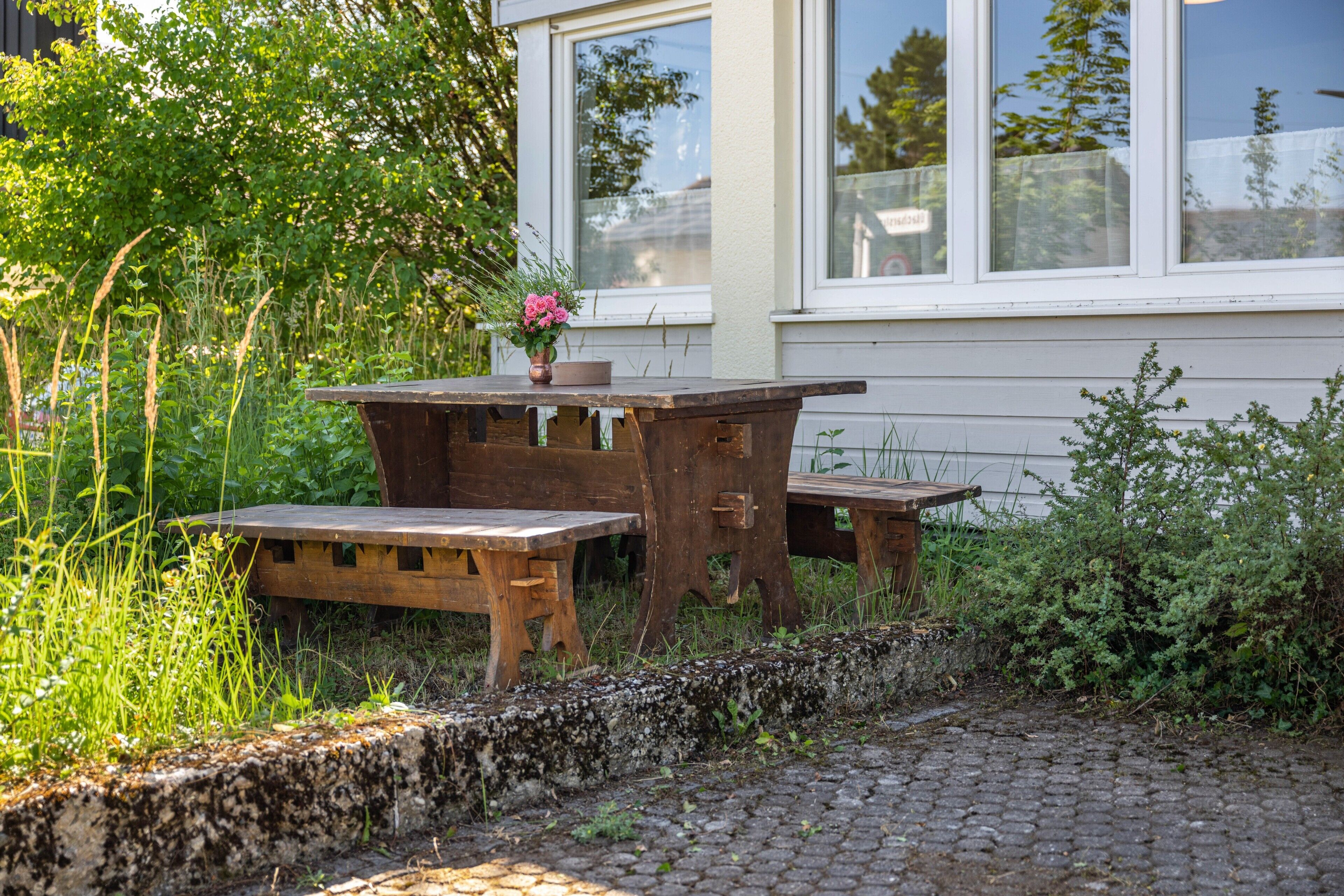 Un espace de restauration extérieur rustique, avec une table et des bancs en bois, entouré de verdure et bordé d’un chemin pavé.