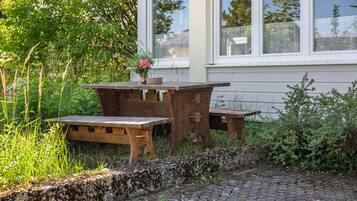 Un espace de restauration extérieur rustique, avec une table et des bancs en bois, entouré de verdure et bordé d’un chemin pavé.