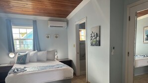 A bedroom with a bed, a nightstand, a wall-mounted air conditioner, a door, and a window with curtains. - BEACHSIDE TWO BEDROOM VILLA - OCEANVIEW (Georgetown)
