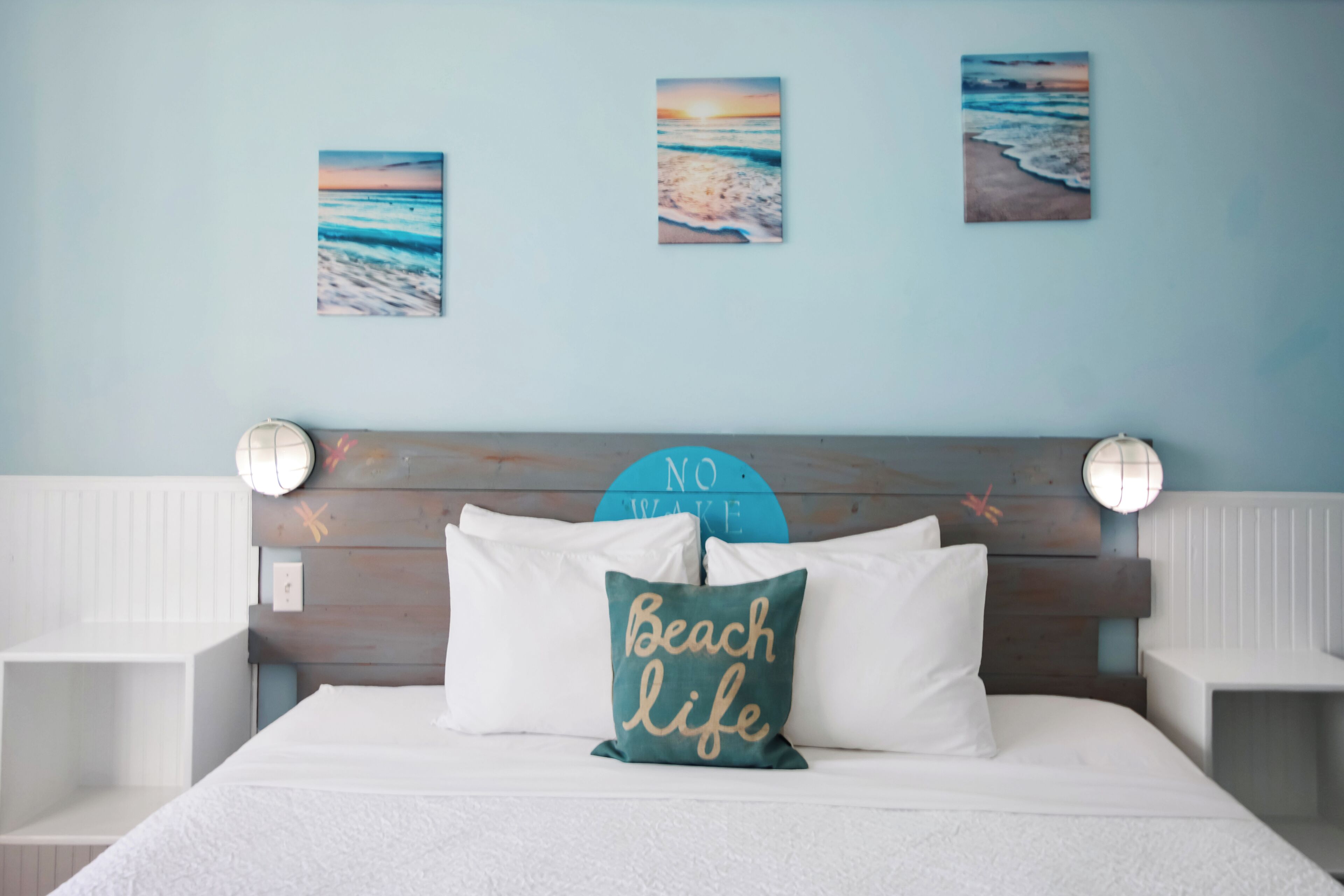 A bed with white bedding, a 'Beach life' pillow, and three beach-themed wall art pieces.