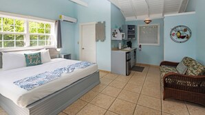 Een slaapkamer met een bed, een kleine kitchenette, een rieten bank en een raam met uitzicht op groen.