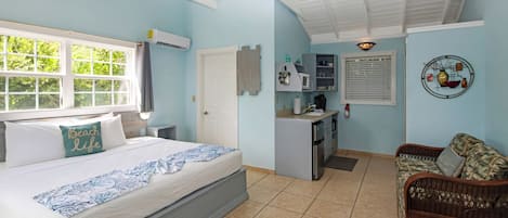 A bedroom with a bed, a small kitchenette, a wicker sofa, and a window with a view of greenery.