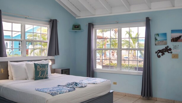 Een slaapkamer met een bed, een plafondventilator, een raam met gordijnen en een muur met een schilderij.