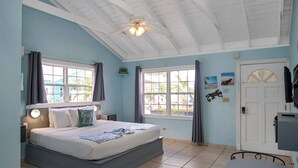 Een slaapkamer met een bed, twee ramen, een plafondventilator, een deur en een klein tafeltje.