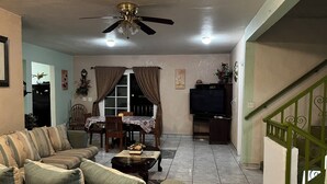 A living room with a sofa, a dining table with chairs, a TV, and a ceiling fan. - 9-Bedroom Ocean View Home in Vista Marina for Large Groups (Vista Marina)