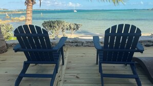 Två Adirondack-stolar står på ett trädäck med utsikt över en strand, en palmtree och havet.