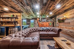 Ein rustikales Blockhäuschen mit einer ledernen Eckcouch, Holzwänden, einer Bar mit Flaschen und Gläsern und einem schönen Ausblick durch eine Tür.